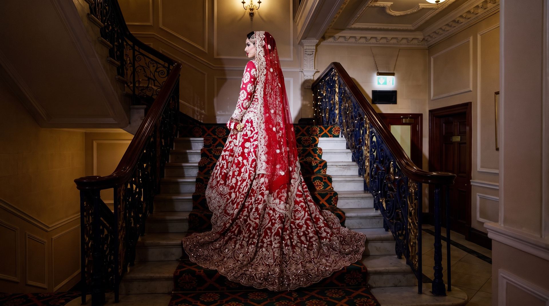 A bride in a deep red bridal lehenga descending a grand staircase, the embroidered train spread across the steps behind her