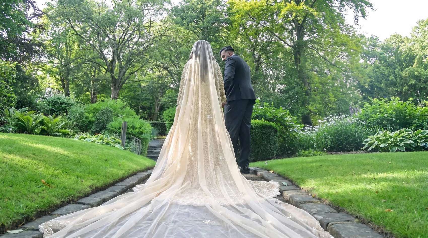 A bride and groom holding hands in a garden, seen from behind, her embroidered veil trailing along the path