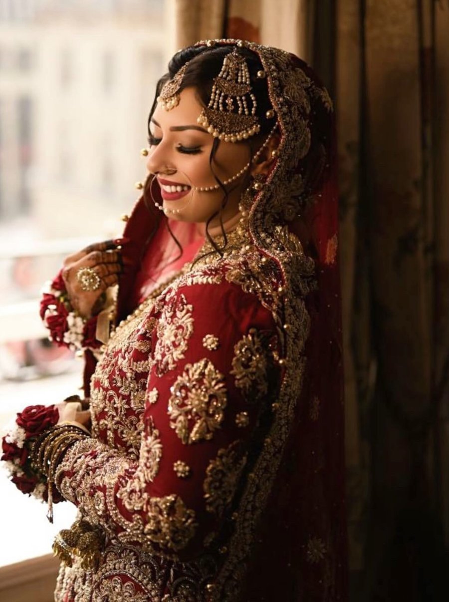 A bride laughing by a window, wearing a red and gold Dulhan Boutique bridal outfit with a rose bouquet in hand