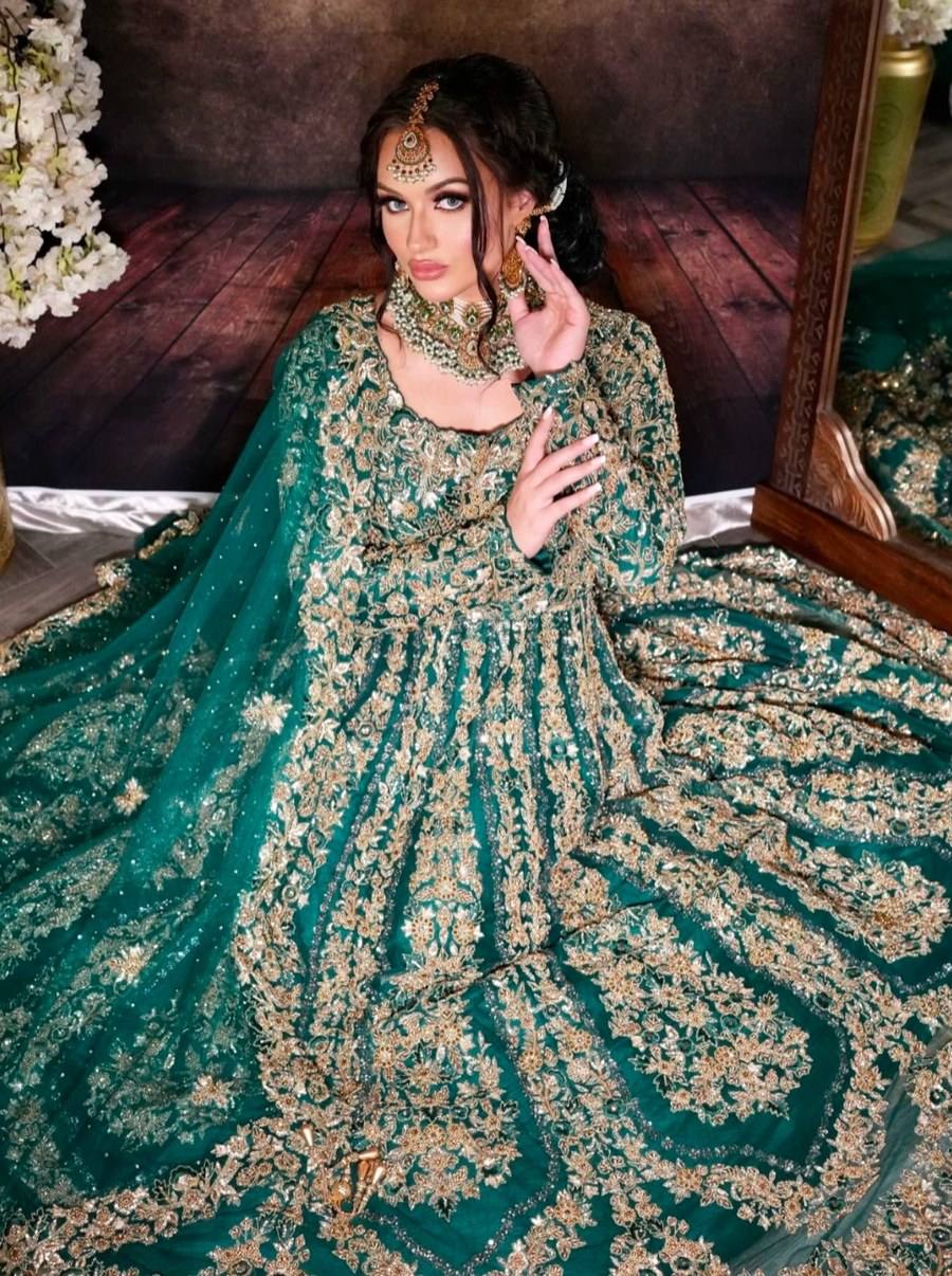 A bride in an emerald green bridal gown with dense gold floral embroidery, kneeling before a mirror with matching jewellery and flowers beside her