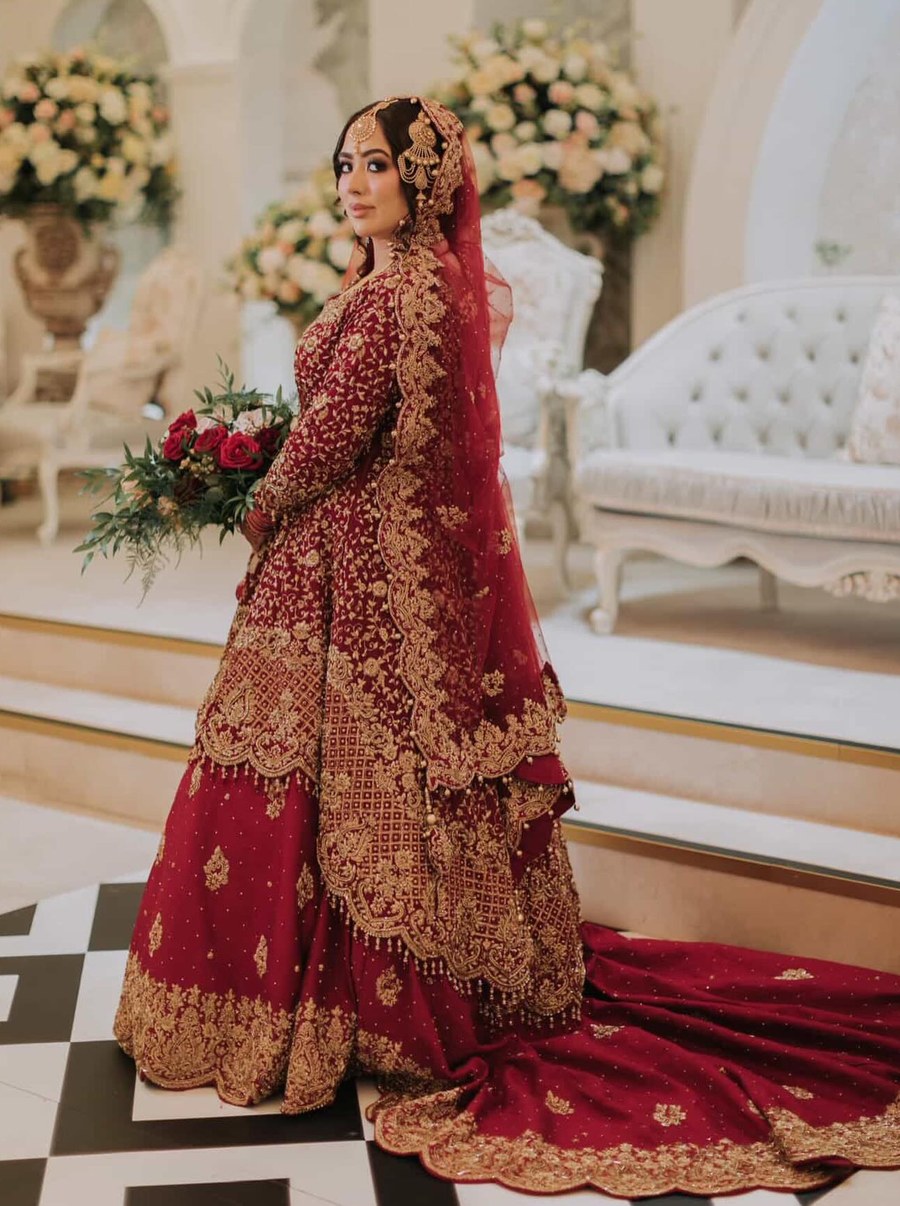 A bride in a deep red bridal lehenga walking with a bouquet of roses, heavy gold embroidery and scalloped tiers trailing into a long train