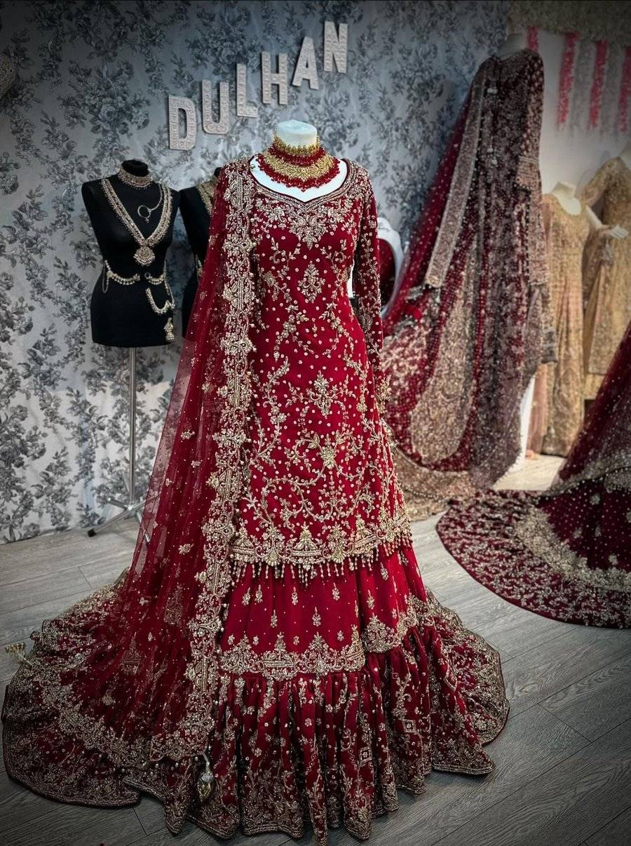 A deep red bridal lehenga on a dress form in the shop, gold floral embroidery and a long train, jewellery on a stand beside it and the DULHAN letters on the wall behind